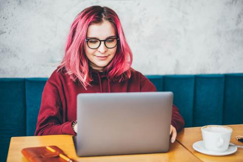 Junge Frau mit pinken Haaren und Brille arbeitet am Laptop.