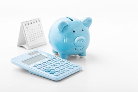Blaues Sparschwein steht neben blauem Taschenrechner und vor einem Kalender