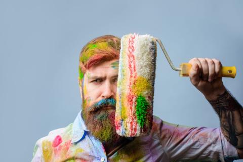 Farbbekleckster Mann hält sich eine bunte Farbrolle vors Gesicht