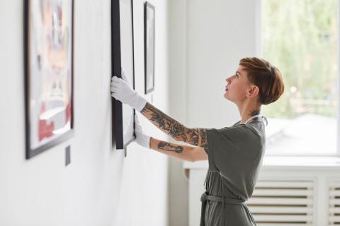 Frau mit Tattoo-Sleeve hängt in einem Museum ein gerahmtes Bild an die Wand