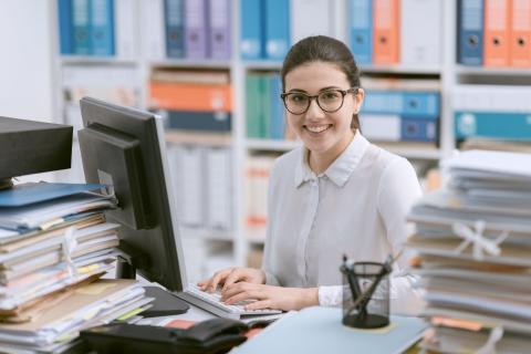 Junge Beamtin arbeitet am Schreibtisch umgeben von Aktenbergen