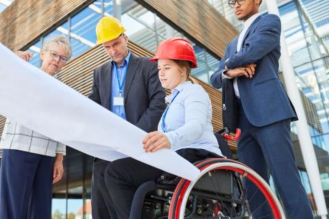 Junge Ingenieurin im Rollstuhl erläutert auf einer Baustelle Pläne