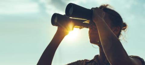 Person steht mit Fernglas vor den Augen vor türkisem Himmel
