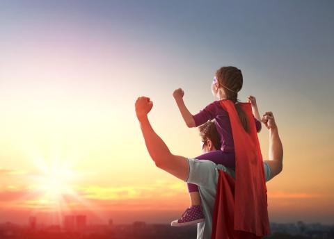 Vater trägt seine Tochter auf den Schultern und beide tragen Superhelden-Capes